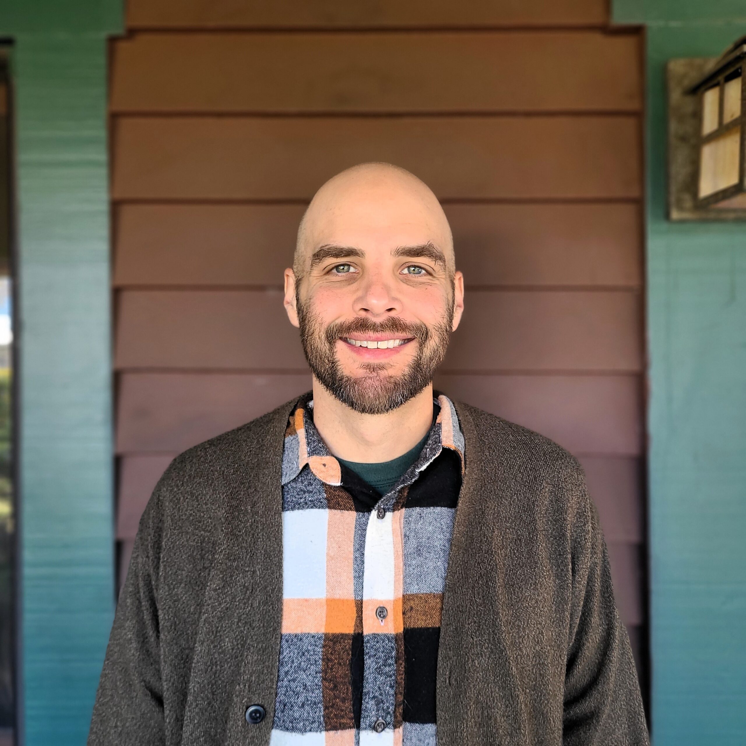 Wayland McQueen wearing an orange, white and black plaid button-up shirt and brown cardigan over an emerald tshirt in front of a brown and green wall