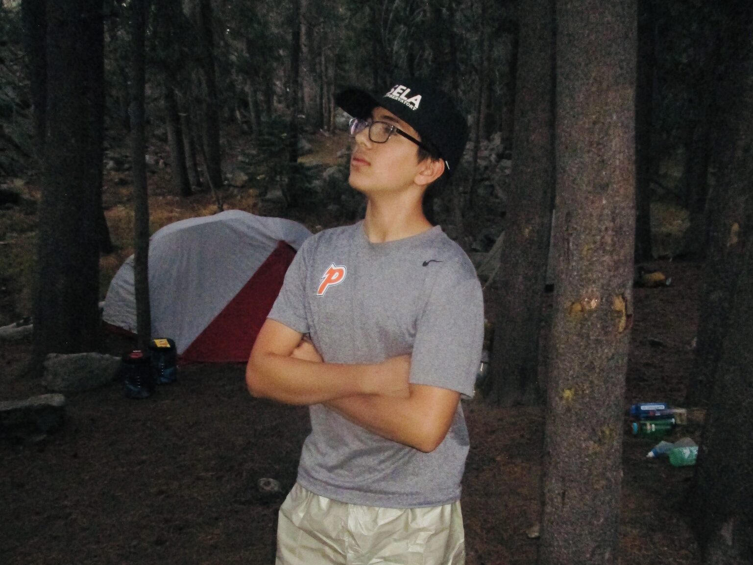 Santiago in a gray t-shirt and black baseball cap with his arms crossed, looking up in the middle of the woods by a tent