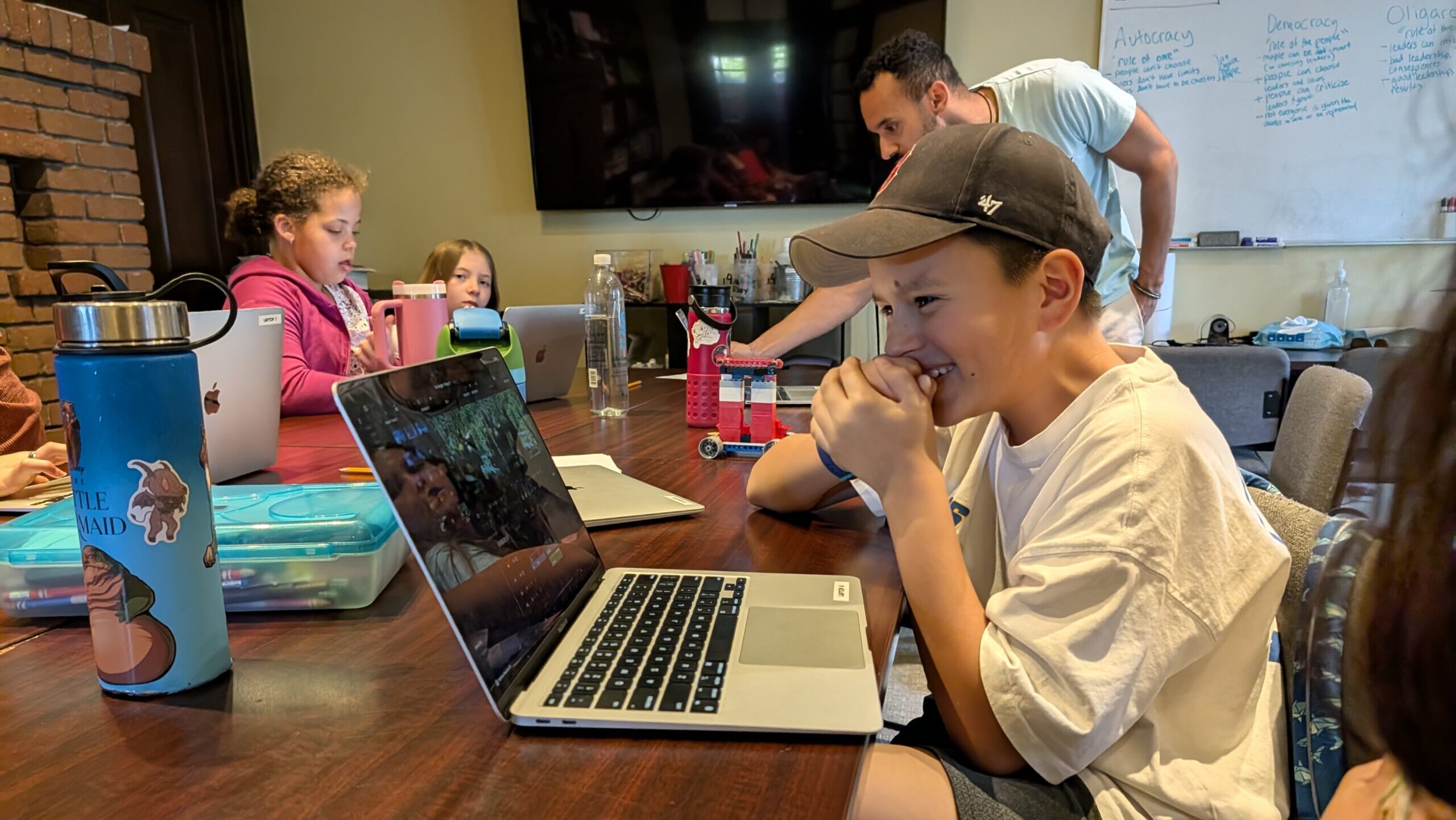 Student in a baseball cap with his hands over his mouth, laughing at his laptop screen while video editing in at IEA's Learning Center