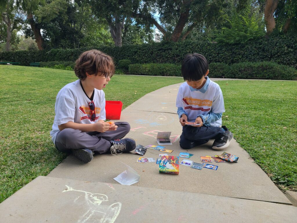 two kids playing with chalk and Pokemon cards on a sidewalk in a park