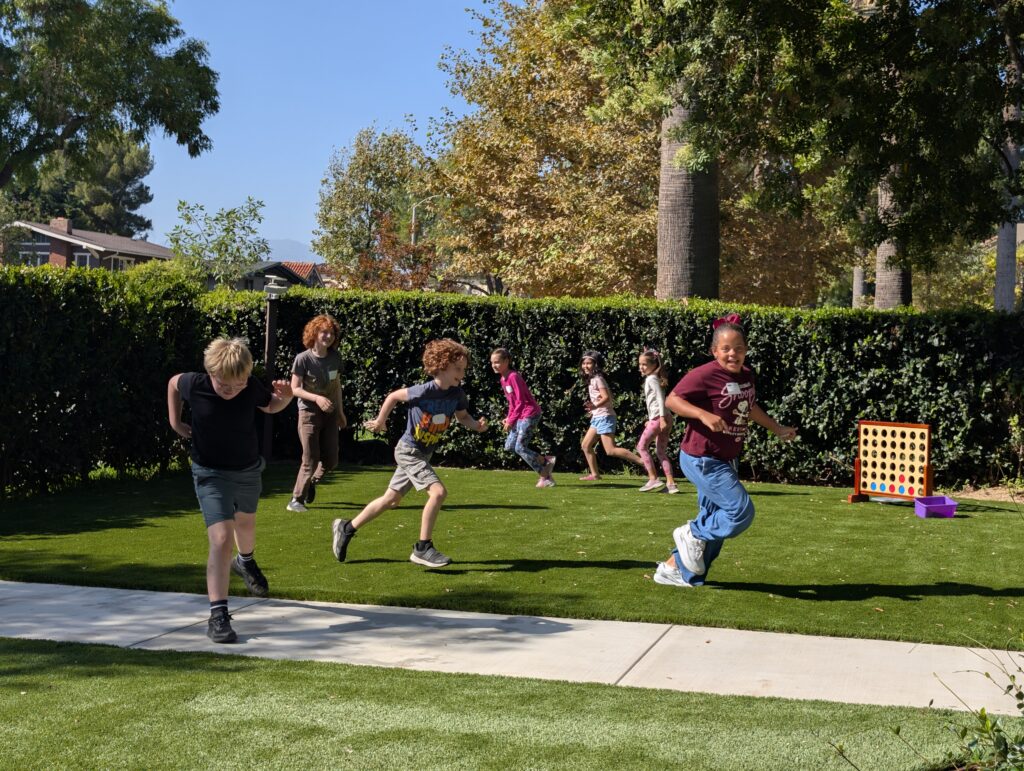 A group of kids playing tag on a lawn near a giant Connect 4 set on a sunny day
