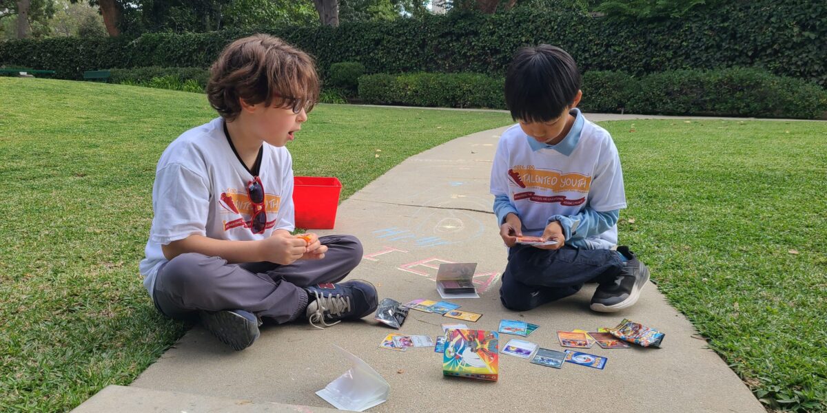 two kids playing with chalk and Pokemon cards on a sidewalk in a park