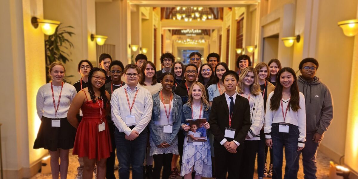 about 30 high school freshmen in dressy-casual attire smiling together, standing in 3 rows in the middle of a hotel lobby with high ceilings