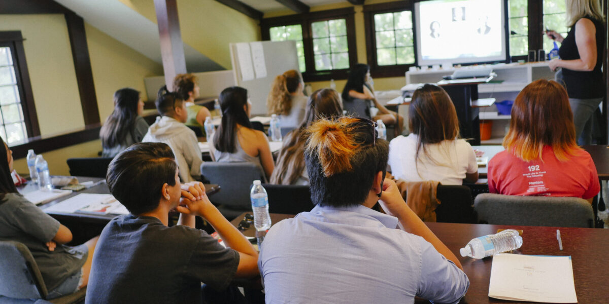 High school students sitting at desks listening to their instructor in front of a monitor