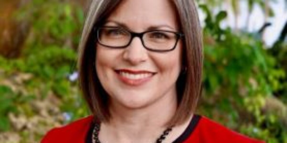 Woman wearing a red jacket and glasses smiling in front of greenery