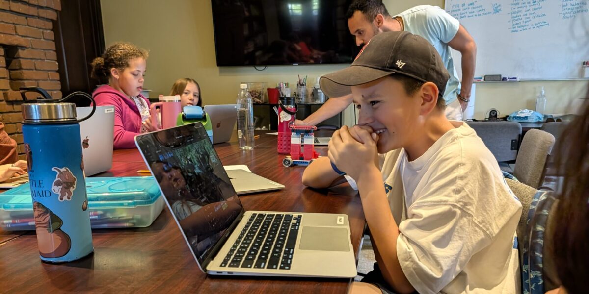 Student in a baseball cap with his hands over his mouth, laughing at his laptop screen while video editing in at IEA's Learning Center